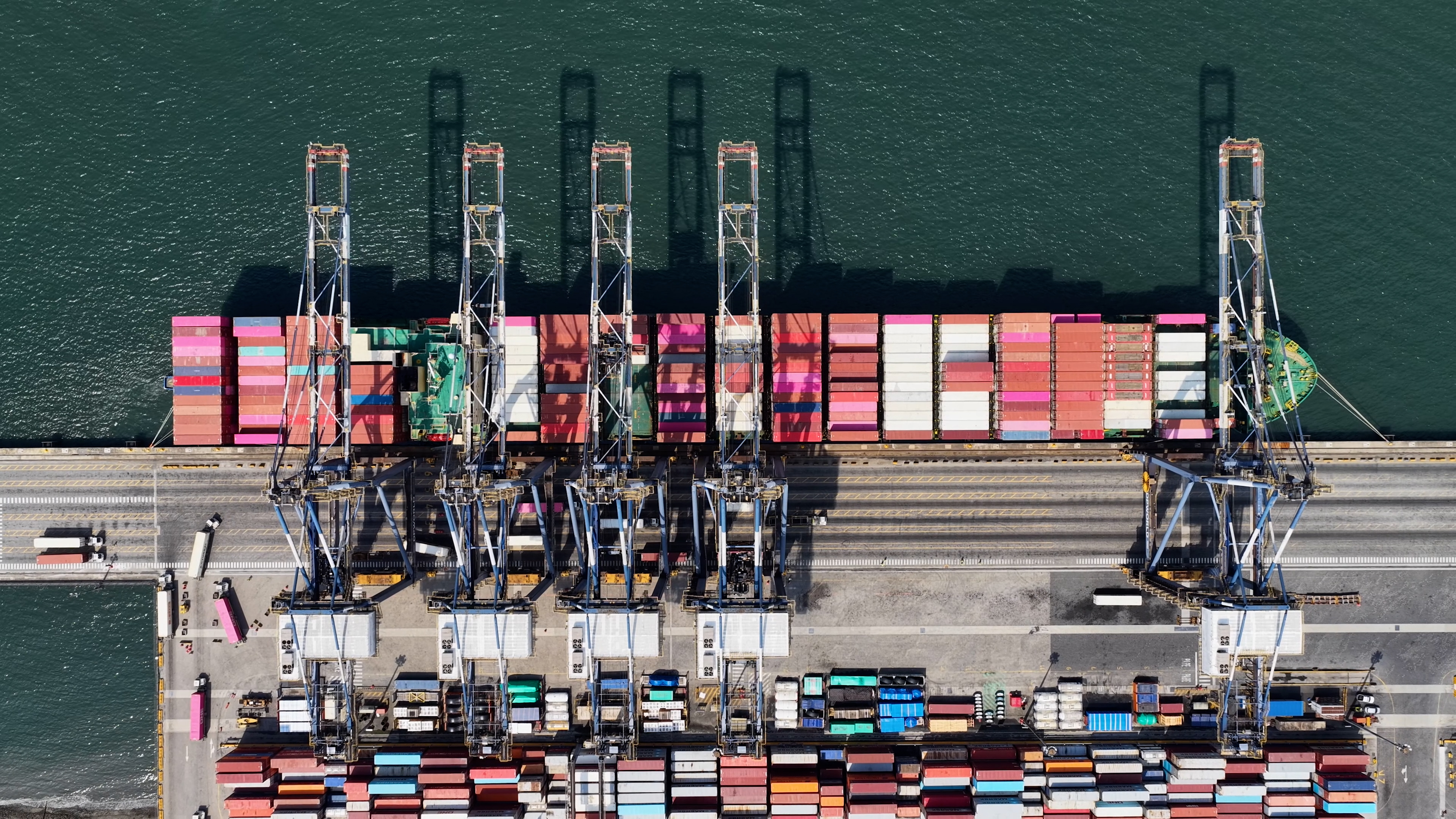 Vista a&eacute;rea de navio cargueiro atracado entre guindastes e cont&ecirc;ineres em terminal portu&aacute;rio